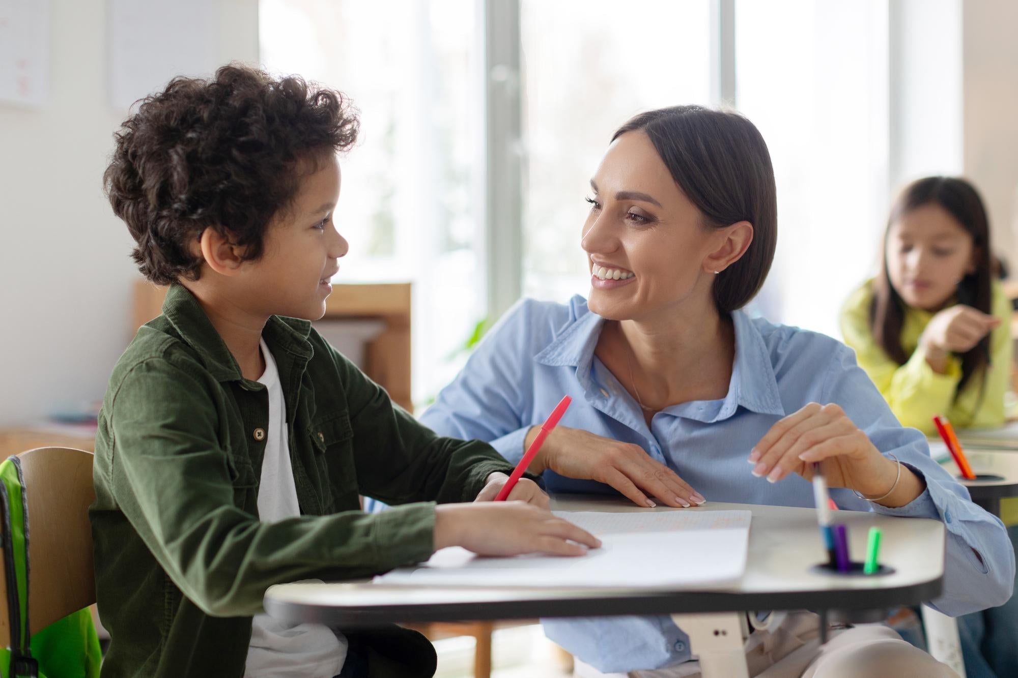 Une AESH souriante accompagne un jeune garçon à son bureau en classe, illustrant l'accompagnement scolaire d'un enfant en situation de handicap