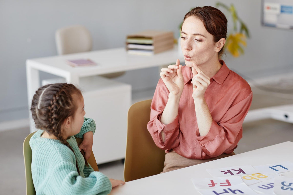 Une orthophoniste travaille l'articulation avec une petite fille à l'aide de cartes-lettres, illustrant une séance de rééducation du langage
